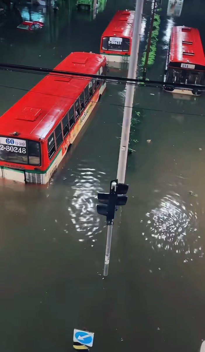 Red public buses with numbers "60 (1-38)" and "2-80248" are stranded in waist-deep floodwaters near a partially submerged traffic light pole, illustrating the severe flooding at Ladprao Police Station and Bang Kapi Market that has made the roads impassable for vehicles.