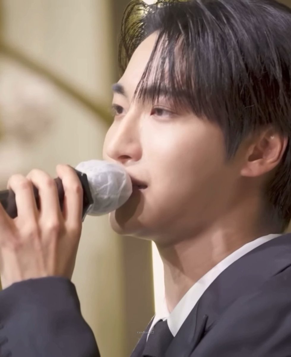 A close-up profile shot of a young man with dark hair singing into a microphone with a white mesh cover, wearing a dark suit and white shirt, with a 'rainbowz22' watermark.