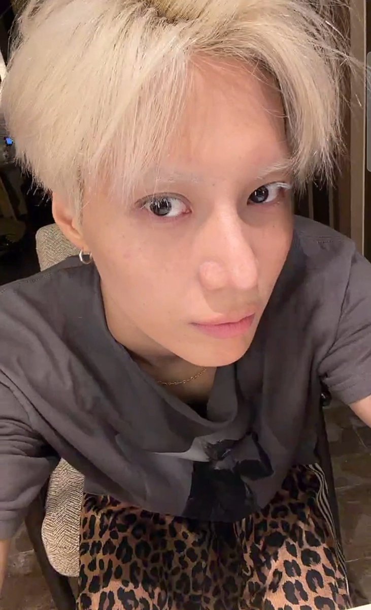 A close-up selfie of a young person with blonde hair wearing a gray top and leopard-print skirt, looking directly at the camera.