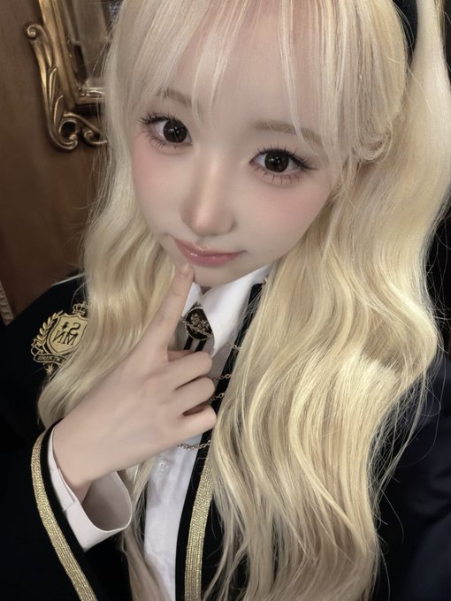A blonde-haired person with large expressive eyes poses with a finger near their lips, wearing a black and white school-style uniform with a crest patch and a striped tie.