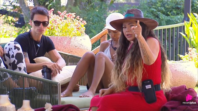 A woman in a red dress and brown hat speaks animatedly while gesturing with her finger, as two other people sit on a couch listening, in an outdoor setting with a 'recordplus' logo visible.
