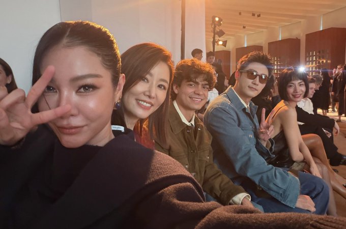 A selfie-style group photo of five people smiling and posing with peace signs at what appears to be an indoor event or venue.