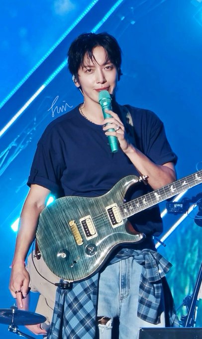 A performer with dark hair sings into a green microphone while playing a teal electric guitar on stage, wearing a navy t-shirt and a plaid shirt tied around his waist.