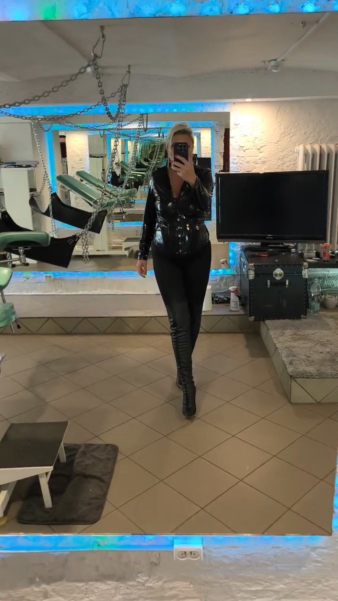 A mirror selfie of a blonde woman wearing a tight black leather outfit and boots, standing in a room with green dental-style chairs, chains hanging from the ceiling, and a TV on a trunk.