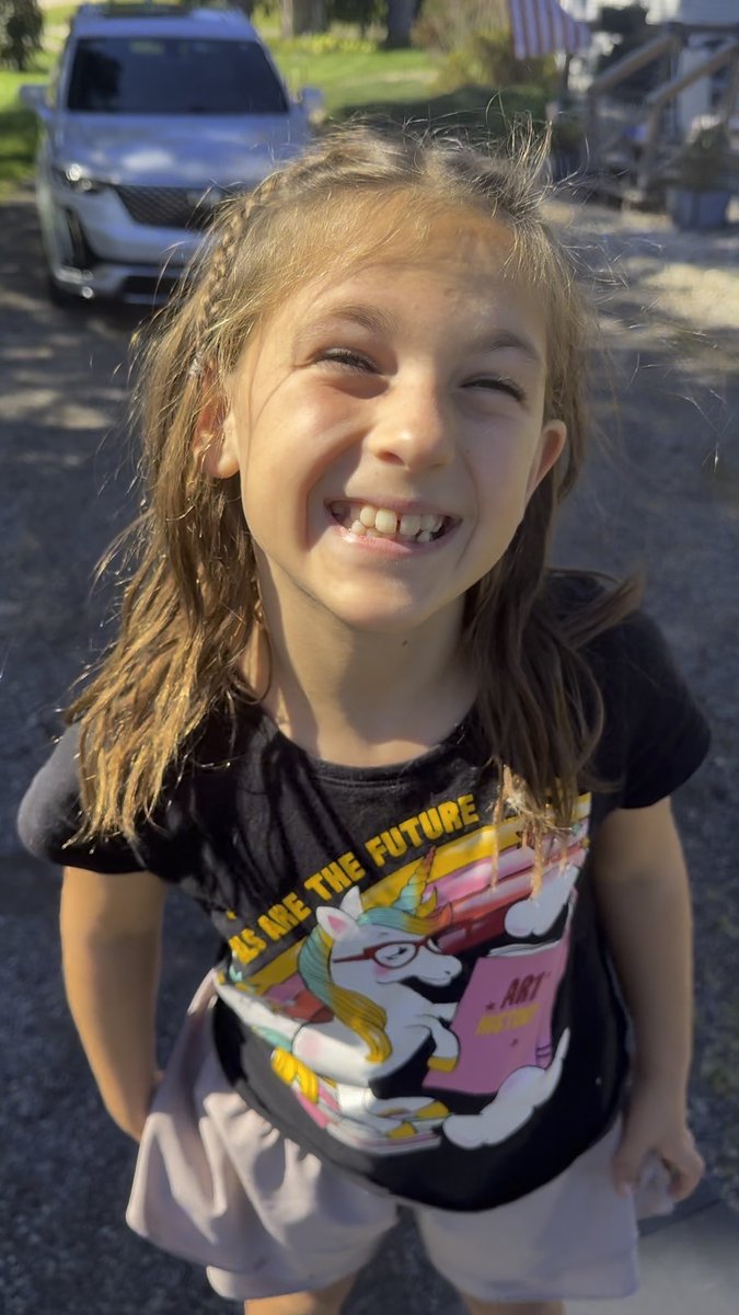 A young girl smiles broadly with squinted eyes outdoors, wearing a black unicorn-themed t-shirt with text reading '...LS ARE THE FUTURE' and 'ART' visible.