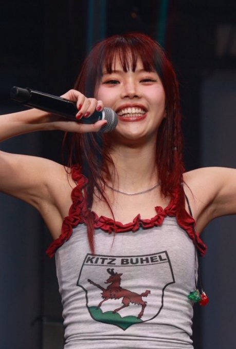 A performer with wet red hair smiles brightly while holding a microphone, wearing a gray tank top with red ruffle straps that displays a 'KITZ BUHEL' emblem featuring a red deer.