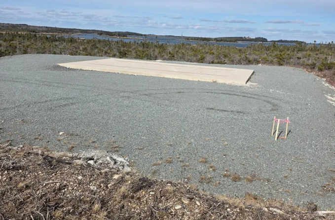 An empty gravel clearing with a concrete slab and survey markers sits in a remote coastal landscape. The image, paired with the post criticizing Carney's $200 million spending to "replace Starlink," ironically illustrates the barren, underutilized result of that investment—no infrastructure or connectivity in sight.