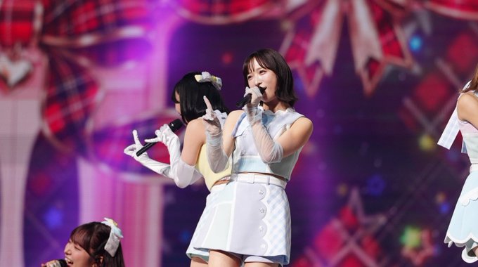 A Japanese idol group performing on stage in coordinated pastel outfits, with the lead singer in a light blue top and white skirt holding a microphone.
