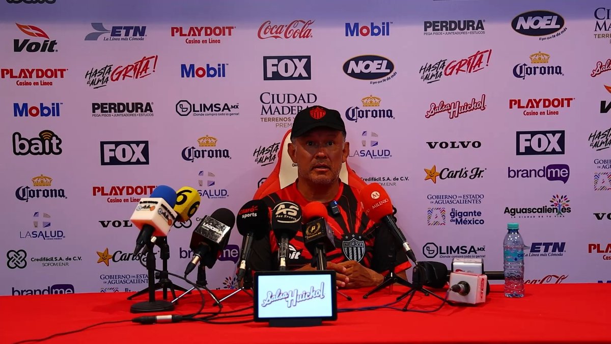 Head coach Juan Reynoso sits at a press conference table wearing a black cap and red-and-black shirt, surrounded by microphones from media outlets like Fox Sports, TUDN, and La Saga. The backdrop is covered in sponsor logos including Coca-Cola, Mobil, Corona, Volvo, and Playdoit Casino en Línea, with a "La Ley y el Orden" placard placed in front of him.