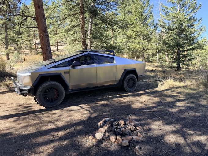 A silver Tesla Cybertruck is parked on a dirt clearing in a pine forest, with a stone-ringed fire pit in the foreground.
