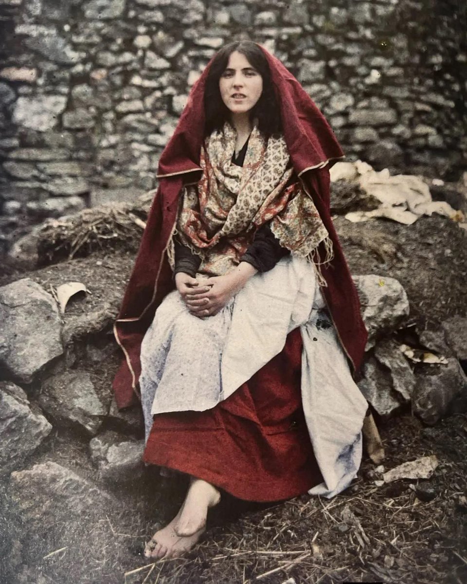 archeohistories's tweet photo. An autochrome color photograph of a young Irish girl in her traditional clothes from the Claddagh in Galway, Ireland, taken on May 26, 1913, by French photographers Marguerite Mespoulet and Madeleine Mignon-Alba.... 

She sits barefoot on the rough earth, framed by a weathered stone wall, her expression steady and self-possessed. Draped in a deep red cloak and what’s likely a Galway shawl—woven in muted florals and worn with layered modesty—she wears the traditional dress of a Claddagh girl, a style once instantly recognizable along the windswept west coast of Ireland.

This image captures more than clothing; it reflects a way of life that has nearly vanished. The Claddagh was once a fiercely independent fishing village just outside Galway City, defined by its tight-knit community, Gaelic language, and deep-rooted traditions. Homes were simple thatched cottages, and daily life revolved around the sea. Men fished in Galway Hookers—sturdy, dark-sailed boats crafted for the Atlantic's wild temper. Women like her maintained the homes, tended to their families, and kept the traditions alive, including the now-iconic Claddagh ring: hands holding a heart beneath a crown, symbolizing friendship, love, and loyalty.

Her shawl, likely woven in the 19th century or early 20th, was both practical and symbolic—a sign of status, identity, and warmth in a damp and wind-bitten landscape. Today, such clothing has nearly disappeared, but in her stillness and strength, we catch a glimpse of a culture quietly proud, richly textured, and deeply feminine.

#archaeohistories