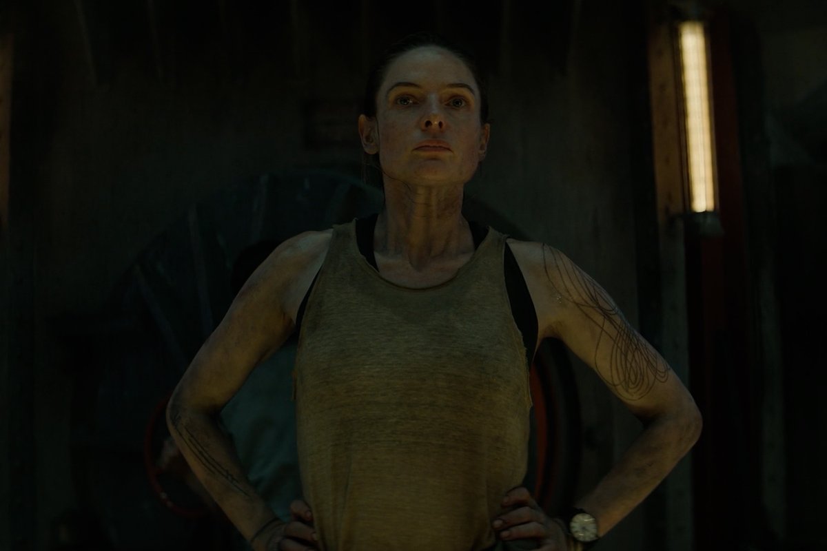 A woman with dirt smudged on her face and body stands with hands on her hips in a dimly lit industrial setting, wearing a tan tank top. The image relates to the post about "Silo" by featuring a determined character from the show in its gritty, dystopian environment.