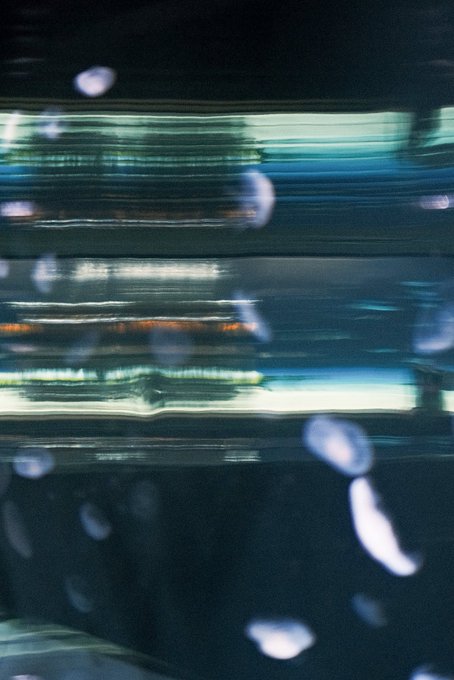 An abstract, blurry image featuring horizontal streaks of green and blue light reflecting on a dark surface, with scattered out-of-focus white light spots resembling bokeh or floating particles.