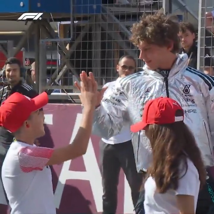 Mercedes driver Kimi Antonelli, wearing a silver AMG team jacket with INEOS and HPE sponsor logos, high-fives a young fan in a white t-shirt and red cap at the F1 Azerbaijan Grand Prix, as another red-capped fan watches nearby with the F1 logo visible in the corner.