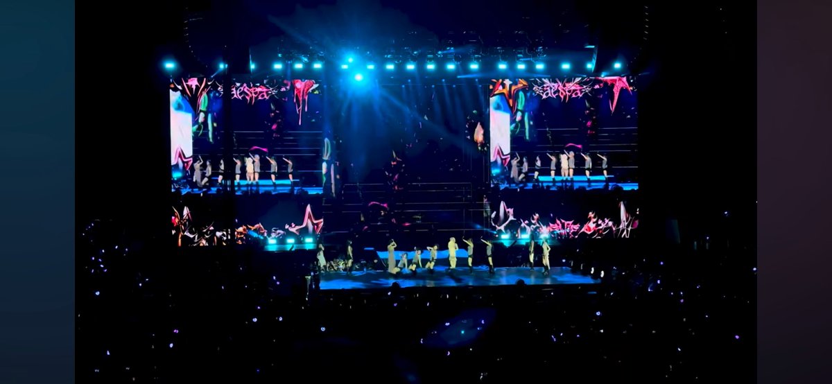 A concert stage is illuminated with bright blue and colorful lights, featuring large LED screens displaying graphics and a group of performers in white outfits on a tiered platform.