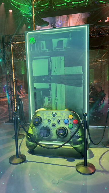 A giant Xbox controller display stands behind velvet ropes, bathed in green lighting that mimics the iconic Xbox console glow. The "XBOX" branding is clearly visible on the controller, reinforcing the showcase theme.