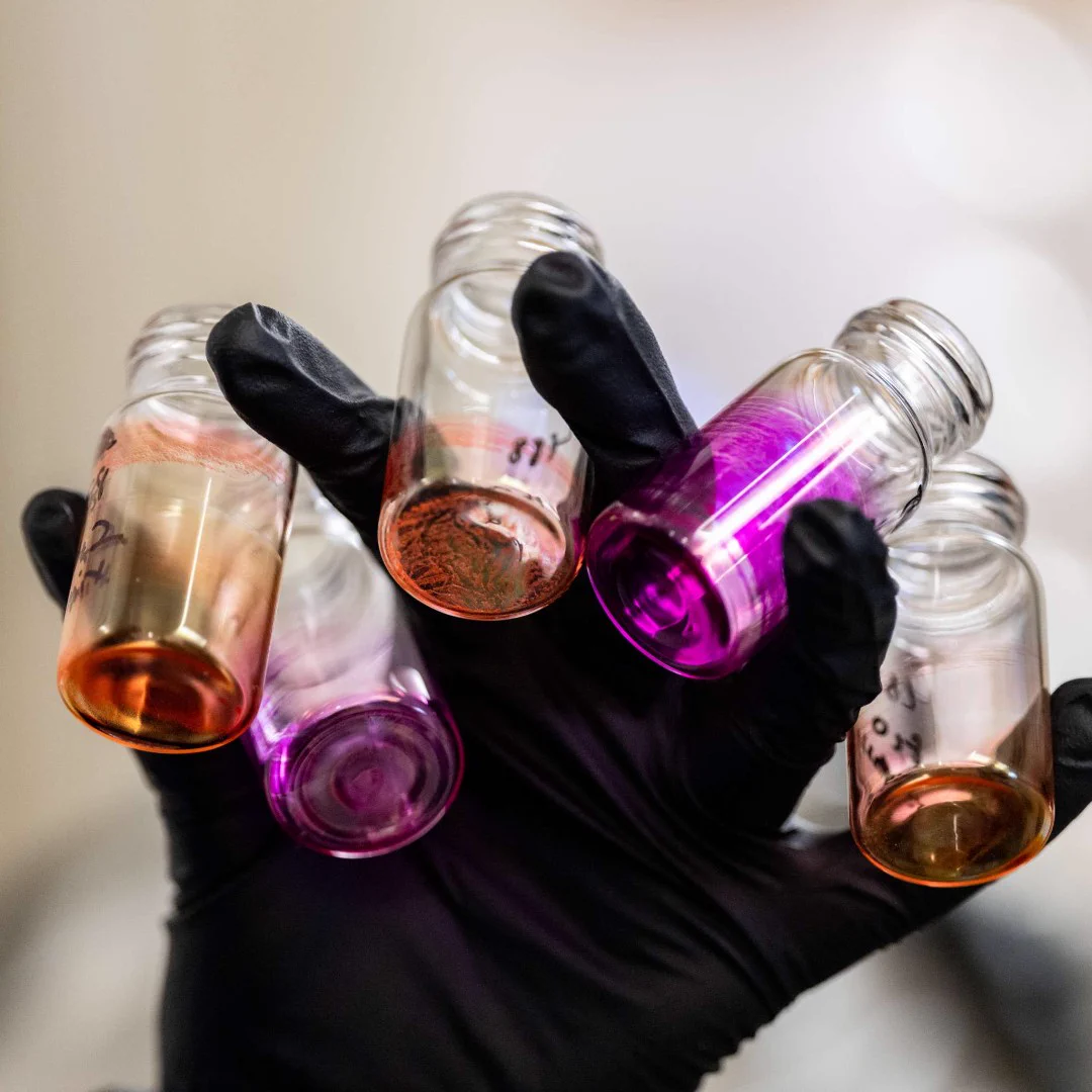 Hand wearing black glove holding five small glass vials with various colored liquids, including pink and orange hues.
