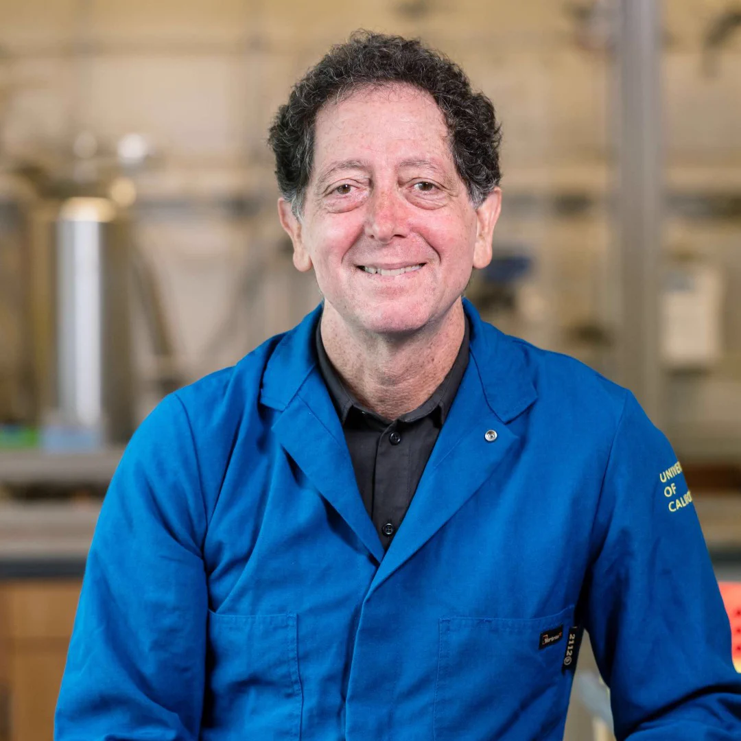 Middle-aged man in a blue lab coat smiling while seated in a scientific laboratory setting.