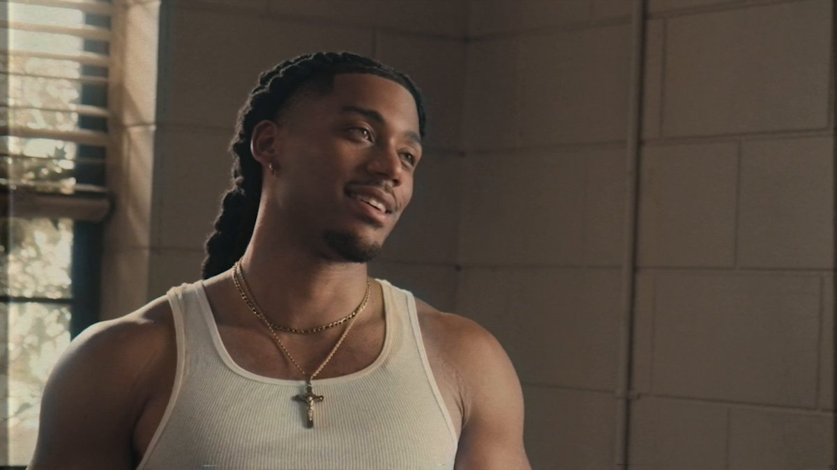 Cornell Young IV as Shaquille in A Different World, shown smiling warmly in a white tank top and cross necklace in a sunlit, tiled interior.
