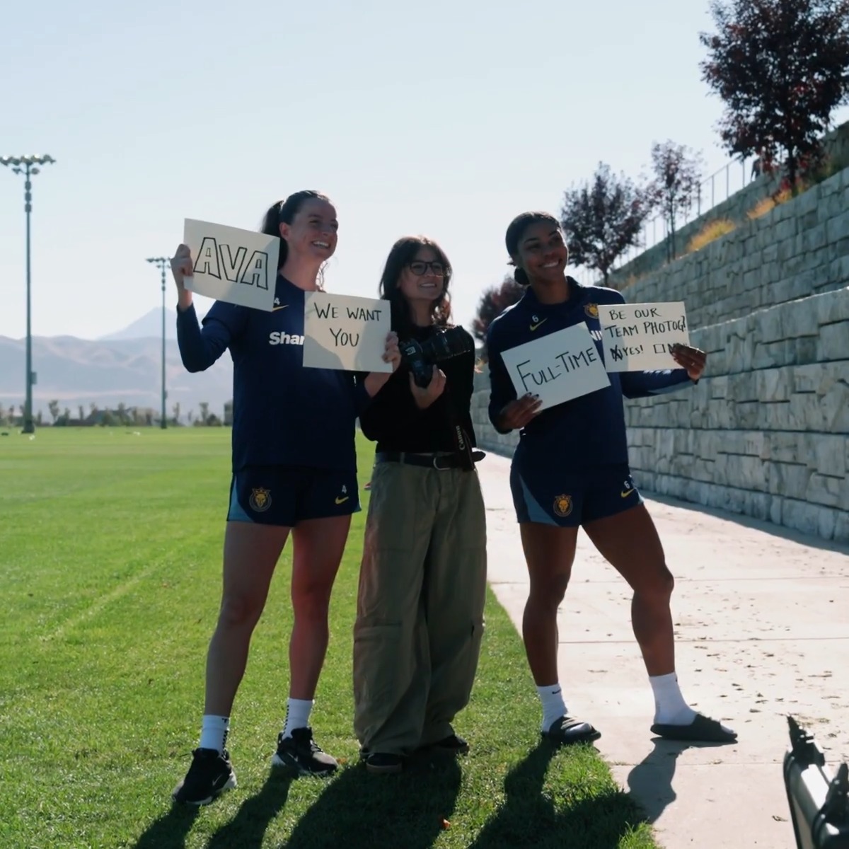 Three young women pose on a grass field; two in matching navy soccer uniforms hold signs reading 'AVA', 'WE WANT YOU', 'FULL-TIME', and 'BE OUR TEAM PHOTOG[ RAPHER]' alongside a friend holding a camera.