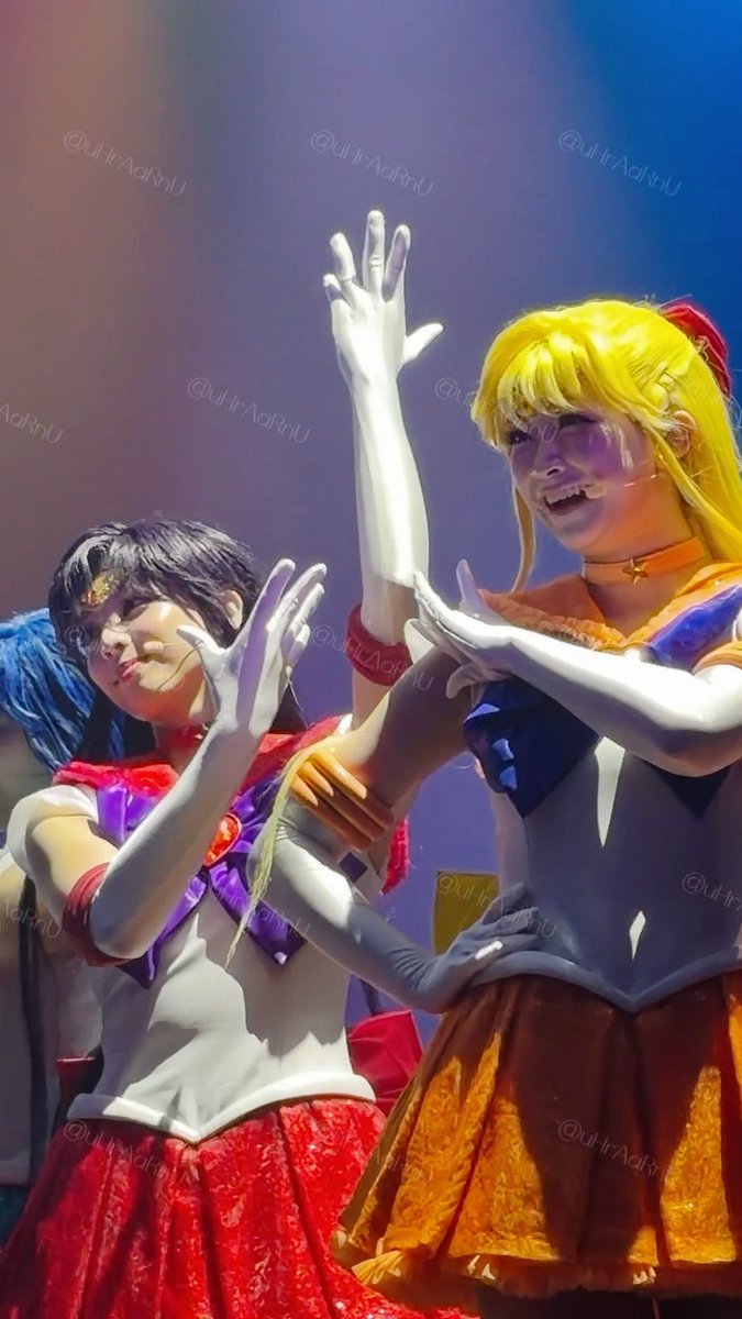 Two cosplayers perform on stage in Sailor Moon costumes, with the character on the right wearing a yellow wig and orange skirt while making a hand gesture, and the character on the left wearing a red skirt and dark wig. The watermark '@UltraAdRnU' appears throughout the image.