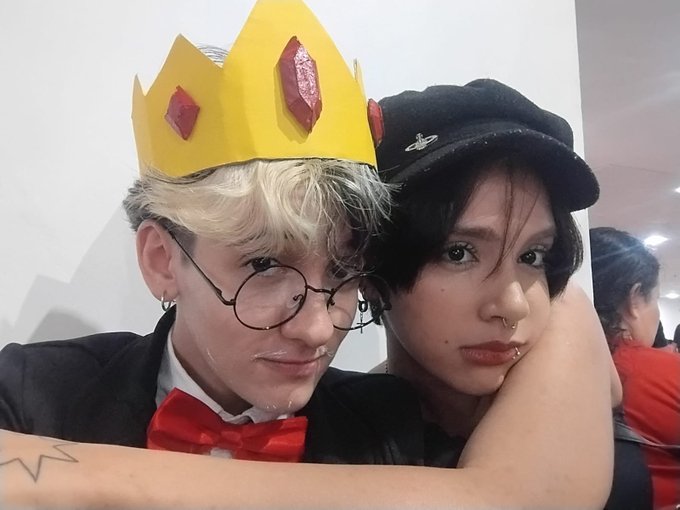 Two young men take a selfie at a costume event, one wearing a yellow paper crown, glasses, and a red bow tie, while the other wears a black cap and has facial piercings.