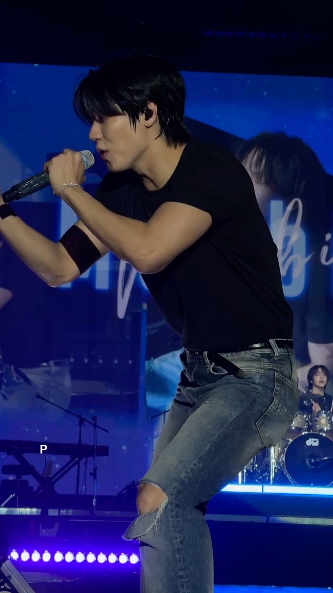A young male performer in a black t-shirt and ripped jeans sings passionately into a microphone on stage, with a drummer visible in the background under blue lighting.