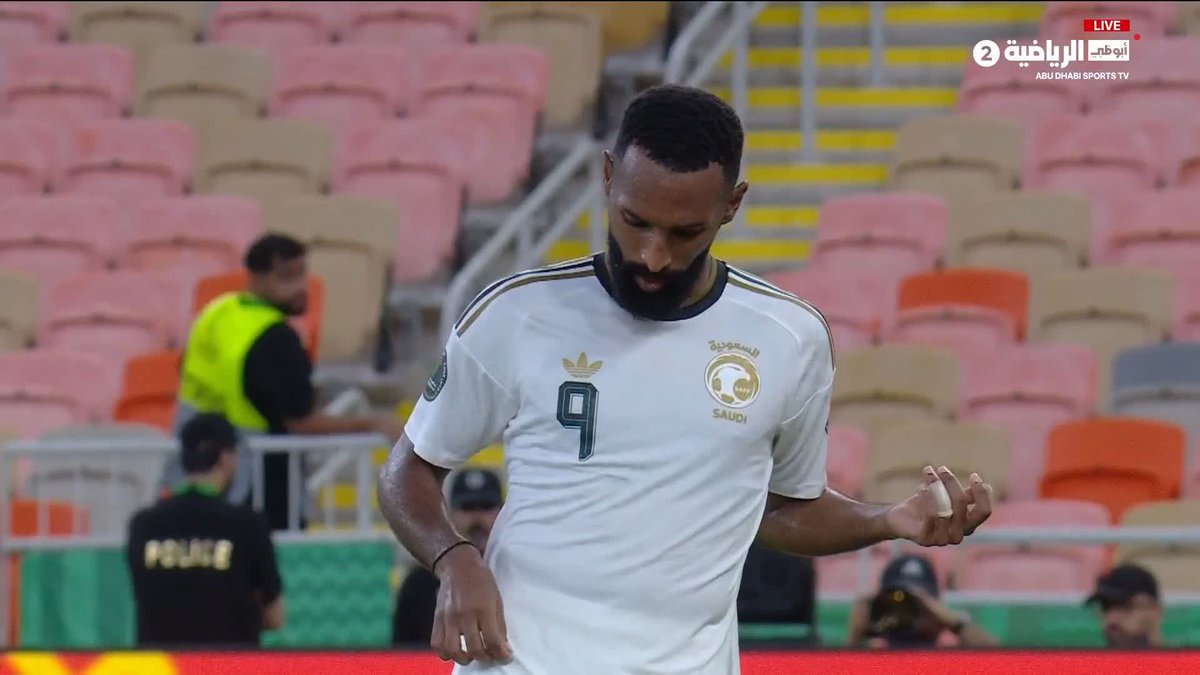 A Saudi Arabia national football team player wearing jersey number 9, representing Firas Al-Buraikan, looks down while walking on the pitch during a match against Oman, broadcast live on Abu Dhabi Sports TV channel 2. The empty stadium seats and visible security personnel suggest this moment captures a quiet pre-match or break in play during the historic Saudi Arabia vs. Oman fixture.
