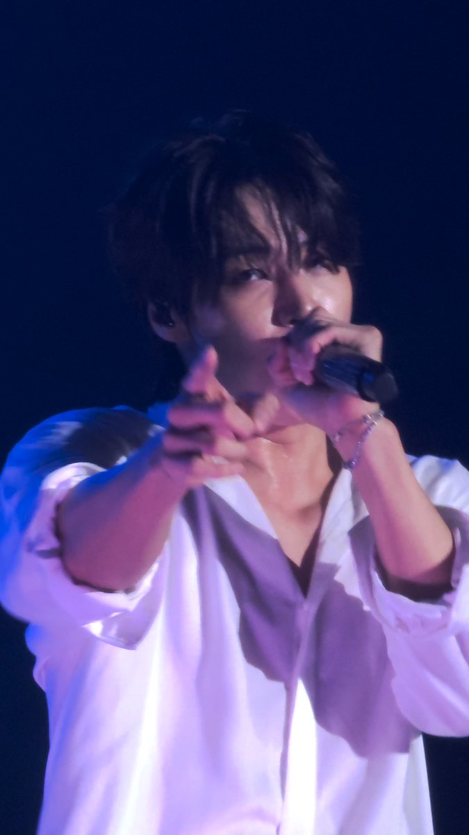 A male performer on a dimly lit stage sings into a microphone while pointing toward the audience, wearing a white button-up shirt and a silver bracelet.