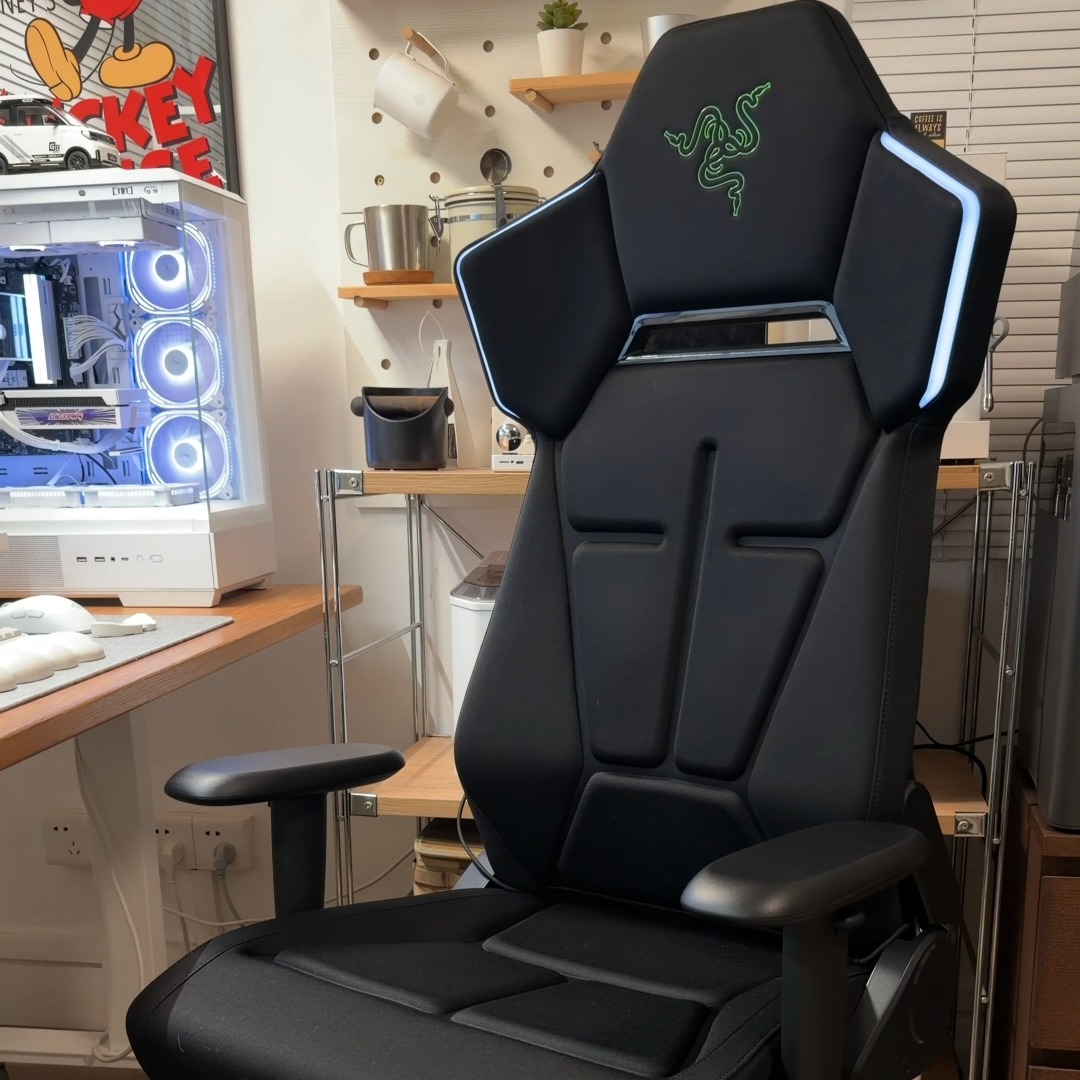 A black Razer gaming chair featuring the iconic green Razer logo sits prominently in front of a desktop setup with an RGB-lit PC case, complementing the advertised "Razer Soma Chroma" RGB peripheral that syncs with the user's existing Razer Chroma-enabled gear across 300+ integrated games.