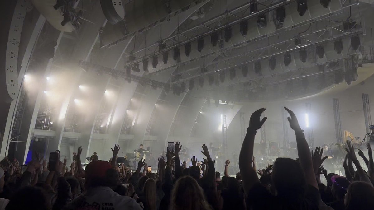 A concert crowd raises their hands at an outdoor amphitheater, with stage lights and smoke creating a hazy atmosphere as performers perform on stage.