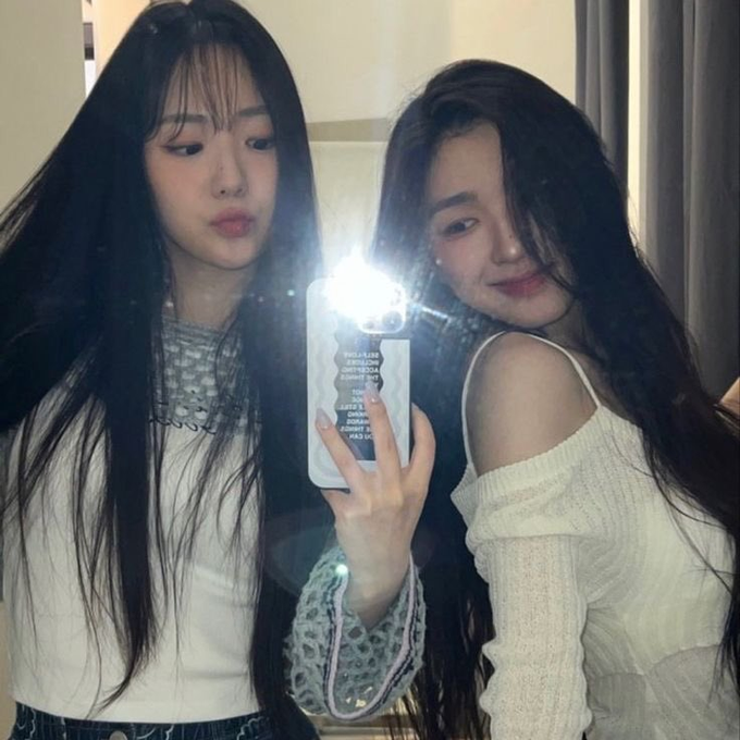 A mirror selfie of two young women with long dark hair, one holding a phone with the flash on, taking a photo together.