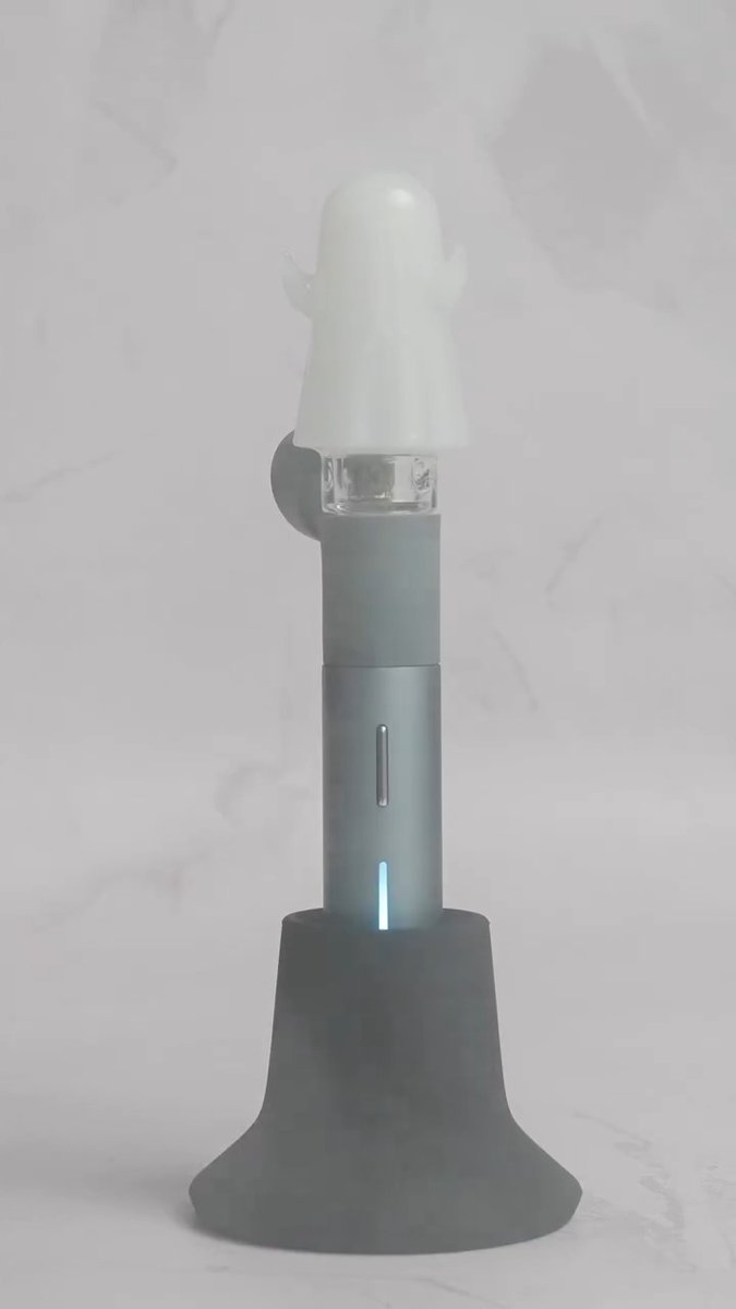A gray adult novelty product with a white silicone top sits on a dark gray flared base with a small blue LED indicator light.