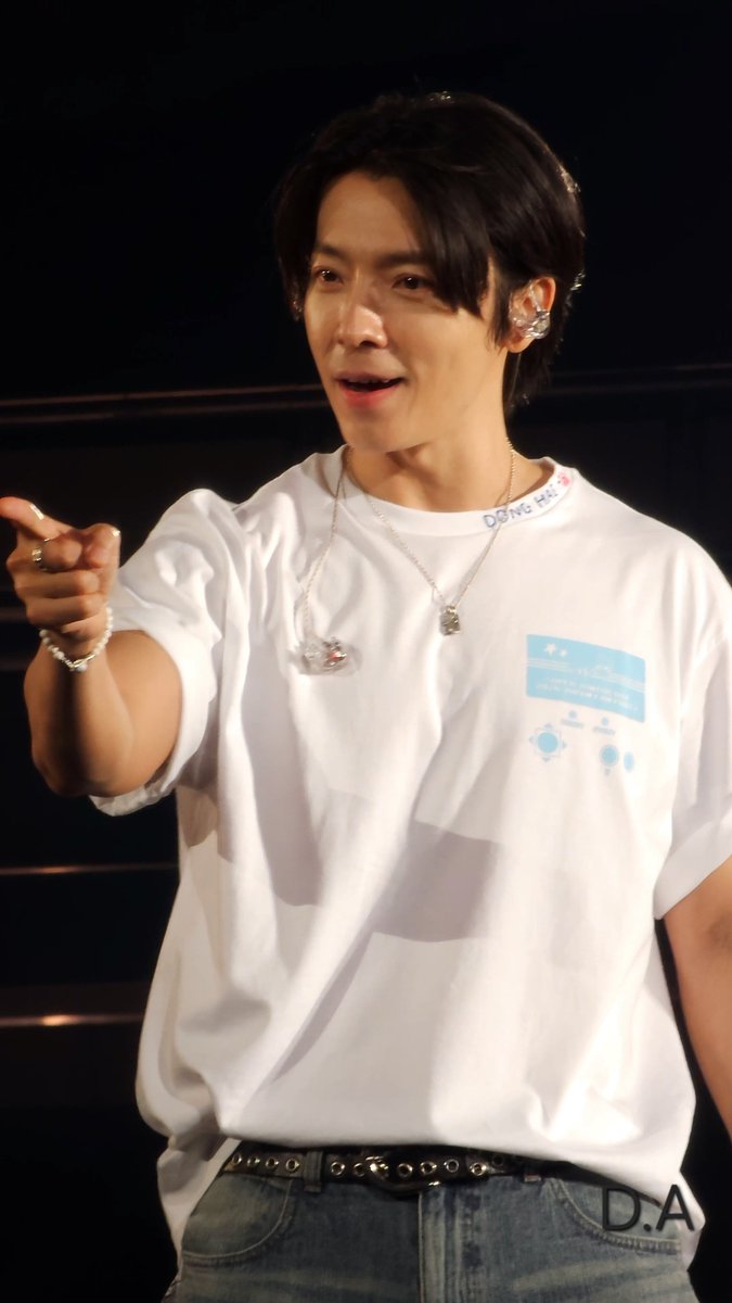 A young man wearing a white t-shirt with text and a graphic, a silver necklace, and an earpiece, pointing to his left with a studded belt visible on his jeans.