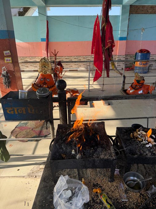 A Hindu temple shrine with a fire pit (havan kund) where flames are burning, flanked by two orange deity figures and red ceremonial flags hanging in the background.