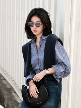 A woman with short dark hair and sunglasses stands outdoors wearing a blue striped shirt under a dark vest, holding a black crescent-shaped handbag.