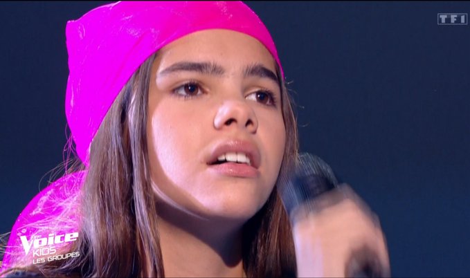 A young performer wearing a bright pink headband sings into a microphone on 'The Voice Kids - Les Groupes' show on TF1.