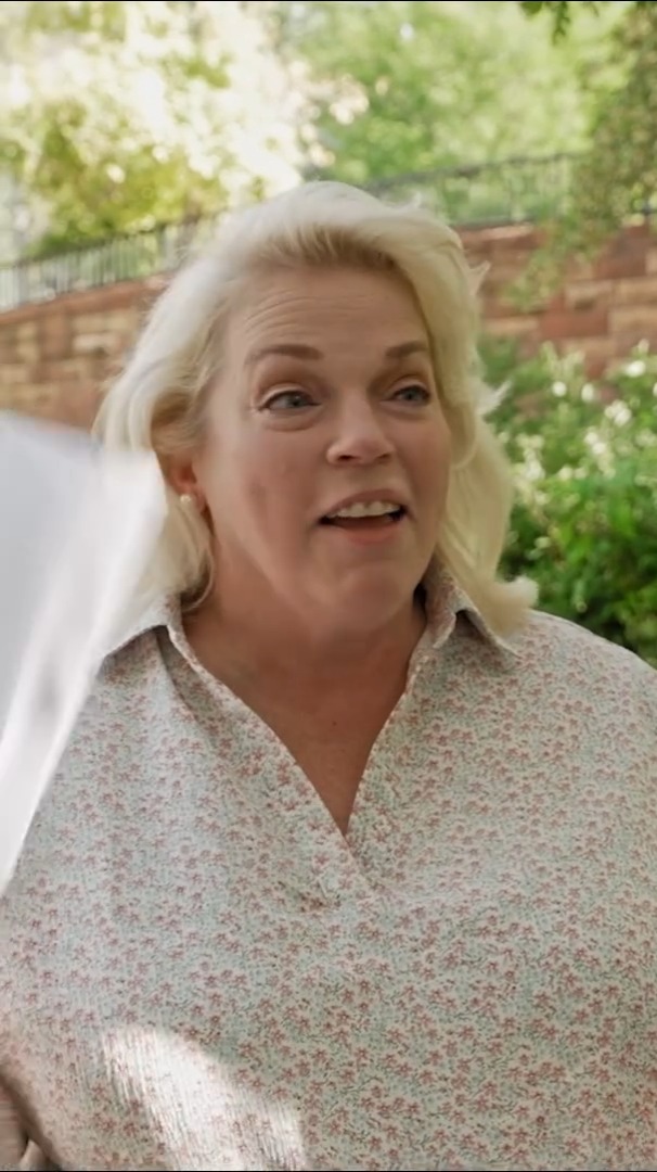 A close-up of a blonde woman outdoors wearing a floral patterned blouse, speaking with an expressive face.