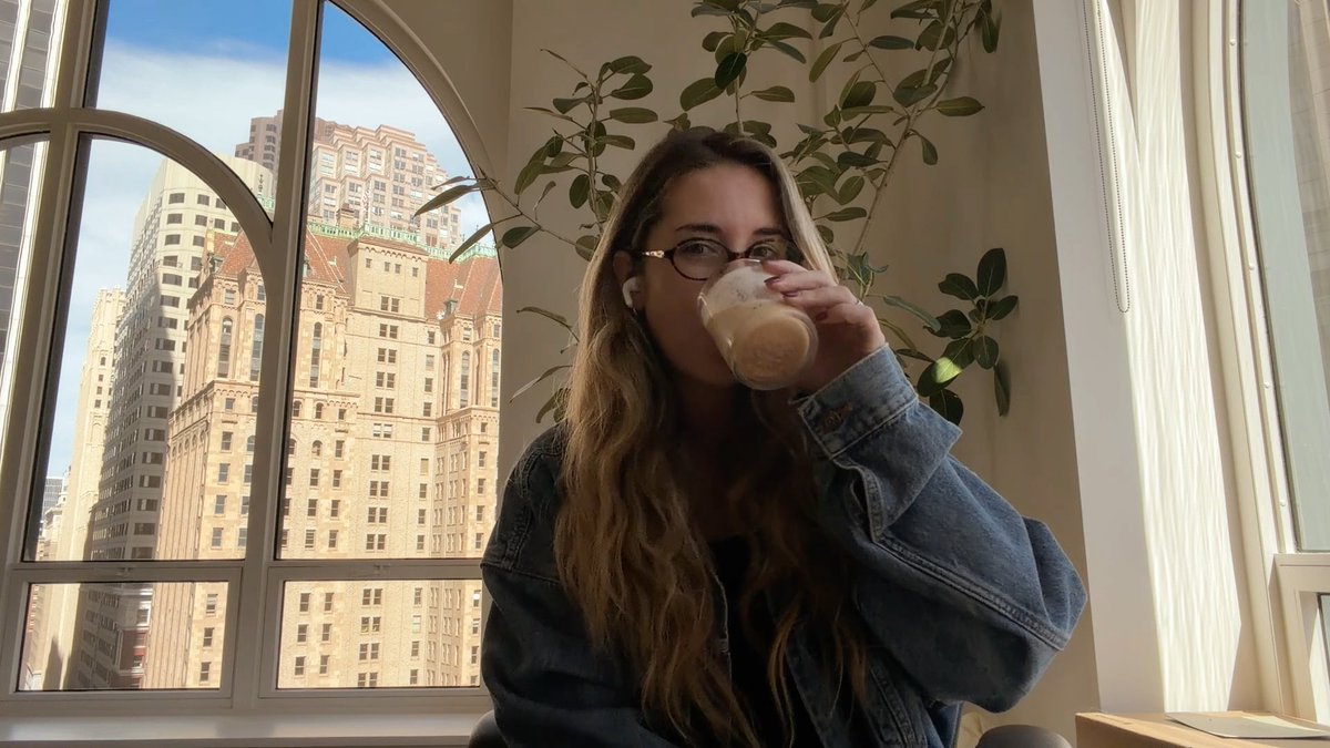 A photograph of a woman with long brown hair and glasses wearing a denim jacket, drinking from a glass of a light-colored beverage while sitting in front of a large arched window overlooking tall city buildings.
