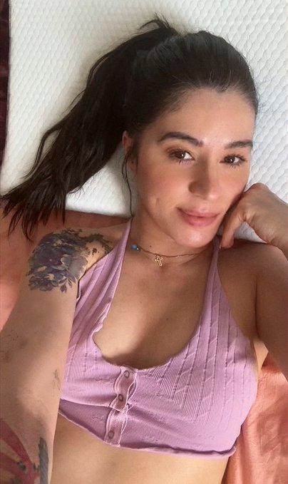 A young woman with dark hair tied back takes a selfie while lying down, wearing a pink ribbed crop top with button details and a small necklace, with a floral tattoo visible on her arm. The image doesn't contain any text, numbers, or game-related visuals, so it appears unrelated to the "9 games that shaped who I am" caption.
