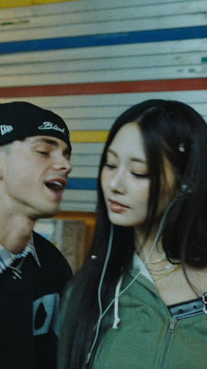 The image shows a young man wearing a black "Blink" baseball cap and a young woman with long dark hair sharing earbuds, posed closely together against a colorful striped wall. This aligns with the post's reference to the song "Blink" and the music video shoot with Tzuyu, with the man's cap prominently displaying the song's title.