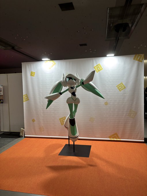 A green and white robotic figure, resembling the Pokémon Gallade or similar character, is displayed on a stand at what appears to be a convention or exhibition, with a white backdrop featuring yellow logos behind it.