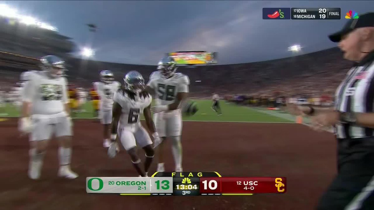 Oregon players #6 and #58 celebrate a touchdown run by Dierre Hill Jr. in the end zone during the 3rd quarter against USC, with the scoreboard showing Oregon leading 13-10 and a flag on the play, while a final score ticker shows Iowa defeating Michigan 20-19.