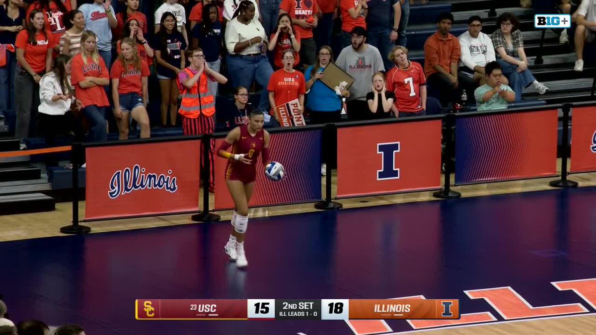 A USC volleyball player in a maroon uniform serves the ball during the 2nd set, with Illinois leading 18-15, as fans watch from the stands behind Illinois-branded barriers.