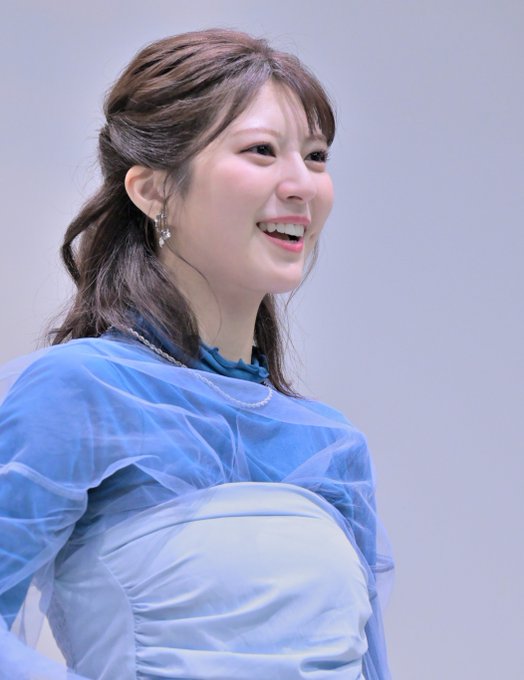 A young woman with brown hair tied back smiles brightly while wearing an elegant blue and white dress with sheer ruffled sleeves.
