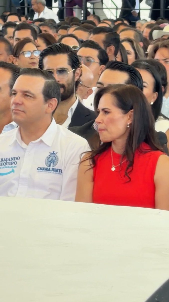 A man wearing a white shirt with 'Guanajuato' and 'Trabajando en Equipo' text sits beside a woman in a red dress at a crowded outdoor event.
