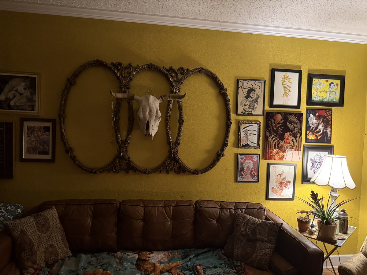 A cozy living room features a yellow wall decorated with an ornate circular frame surrounding a longhorn skull, alongside a gallery wall of various framed artworks above a brown leather couch.