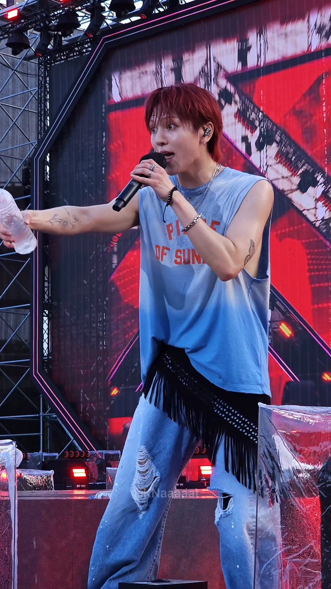 A red-haired male performer wearing a blue sleeveless 'PIECE OF SUN' shirt sings into a microphone on an outdoor stage with a red and black backdrop.