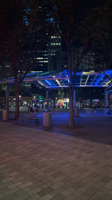 An evening view of an urban plaza featuring a blue-lit pergola structure, trees, benches, and a tall office building in the background.