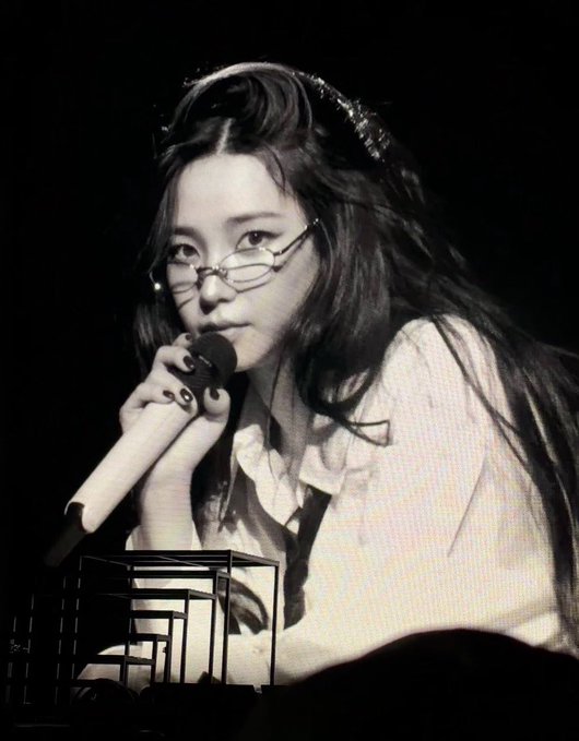 A black-and-white image shows a young woman with long dark hair, wearing glasses, singing into a microphone while a Rubik's cube sits in the foreground.